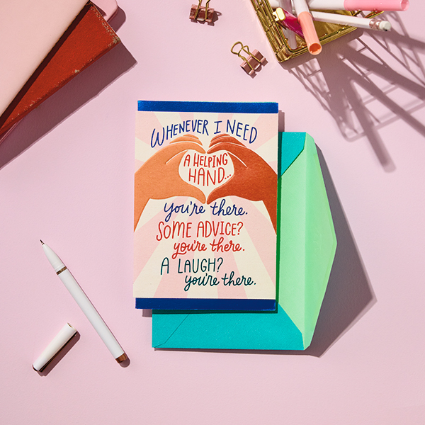 A card meant for a caregiver sits on a light pink tabletop; the front of the card features an illustration of two hands coming together to make a heart shape, and a message that reads, 