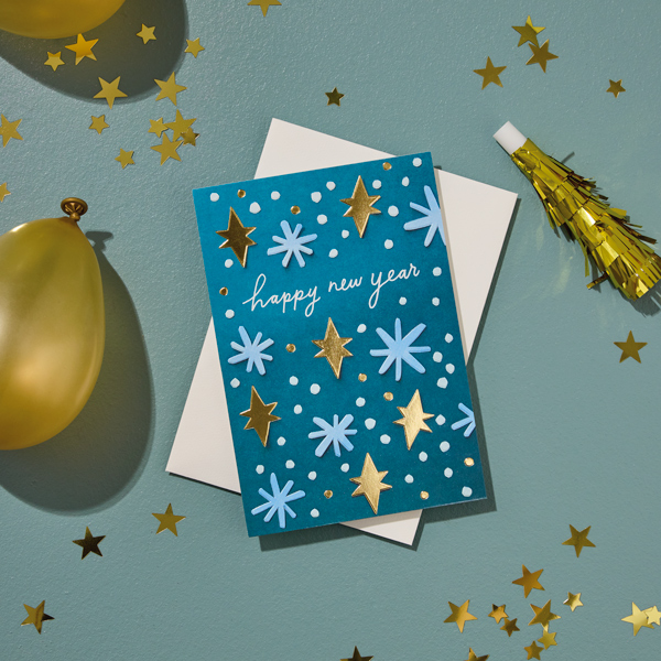 A New Year's card with a blue cover that's covered in gold, light blue and white spangles sits on a gray surface that's scattered with gold star-shaped confetti, gold balloons and a gold party horn.