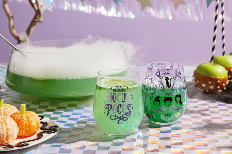 A pair of stemless wine glasses featuring quotes and characters from Disney Hocus Pocus sit on an iridescent checkered tablecloth; the glasses are filled with a green beverage; a large punch bowl filled with the same green beverage sits behind the glasses.