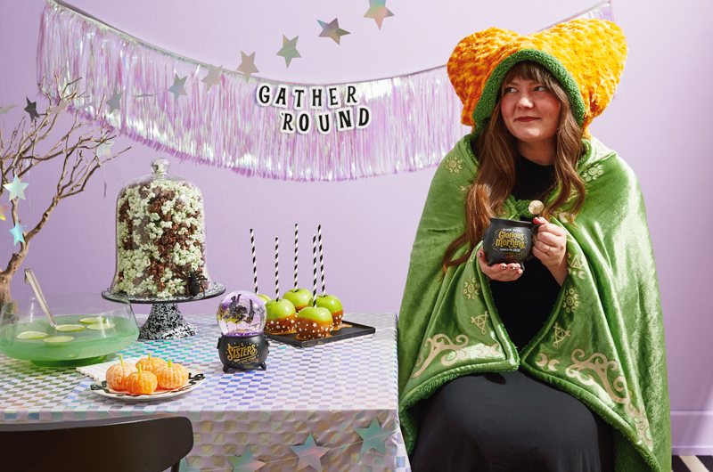 A woman sits in a Winifred Sanderson hooded blanket; the hood of the blanket features bun-like pillows that make the wearer look like their hair is styled like Winifred Sanderson; the woman is holding a mug that reads, 