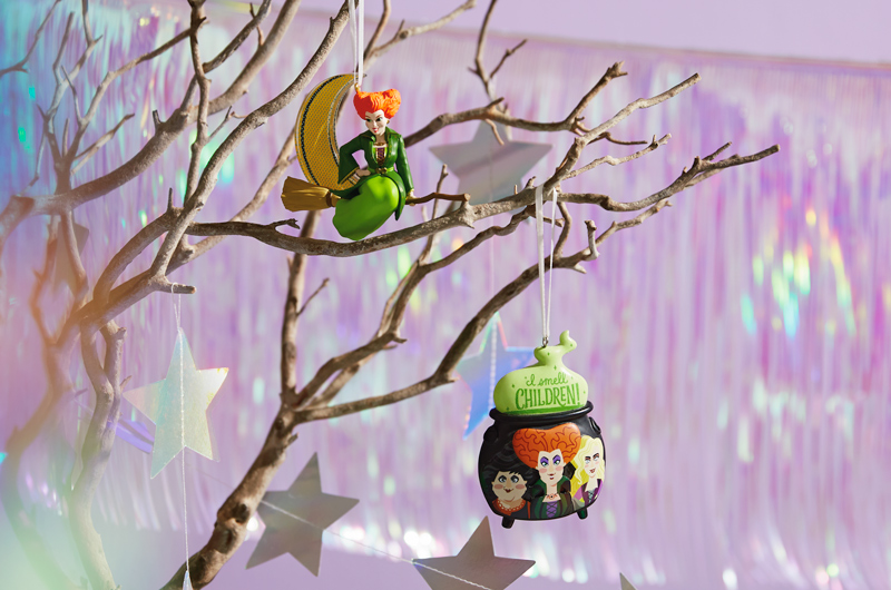 A pair of Keepsake Ornaments featuring Winifred Sanderson and the Sanderson Sisters from Disney Hocus Pocus hang on a bare branch in front of some iridescent hanging streamers.