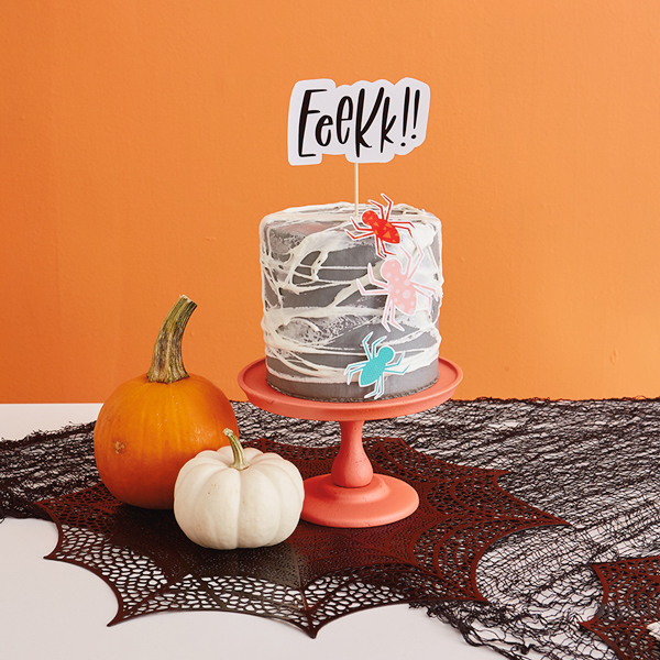 A cake decorated with stretched, melted marshmallow to look like spider webs, paper spider cutouts, and a cake topper on a dowel rod that reads, 