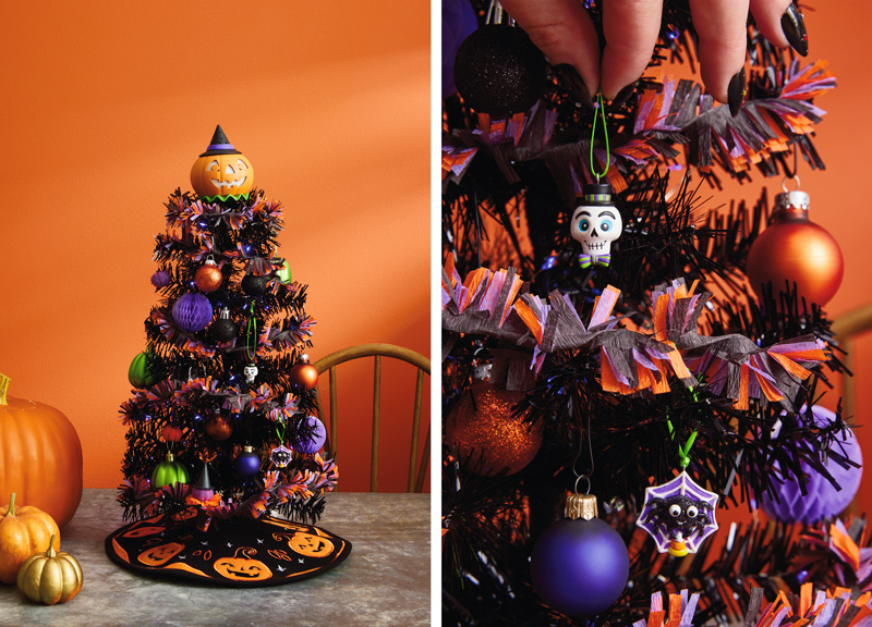 A miniature Halloween tree sits on a gray tabletop next to a trio of different sized orange pumpkins; the Halloween tree is black and is hung with mini ornaments and small glass ball ornaments in shades of purple, green and orange; the tree is topped with a jack-o'-lantern tree topper and has a little black tree skirt embroidered with orange felt jack-o'-lanterns at its base.