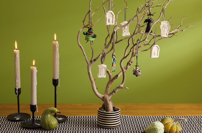 A Halloween tree inspired by Disney Tim Burton's The Nightmare Before Christmas sits in a black and white striped vase on a wooden table, in front of an olive green wall; the tree consists of a bare wooden branch hung with Nightmare Before Christmas Halloween ornaments, and DIY ornaments made from air-dry clay pressed into silicone molds shaped like gravestones.