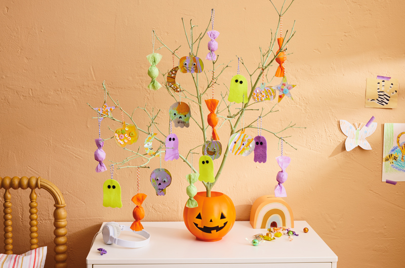 A cute kids Halloween tree consisting of a bare branch spray painted a light green color and hung with DIY ornaments, including neon green ghosts and multicolored skulls, pumpkins, moons and star shapes cut out of chipboard and painted by kids, as well as tissue paper-covered treats hanging from colored string; the branch is set in a classic orange, plastic trick-or-treat pail shaped like a little jack-o'-lantern.