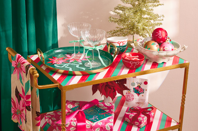 A collection of tableware, wrapped gifts, decor and greeting cards from the Lacey Chabert Collection sits on a gold metallic bar cart that's been decorated for the holiday season.