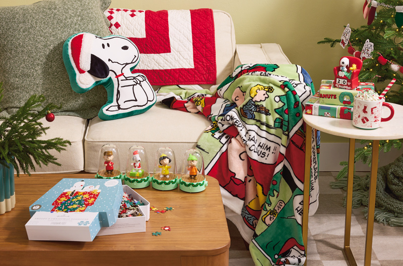 A collection of Peanuts holiday gifts and decor items is scattered around a living room corner that's been decorated for the holiday season.
