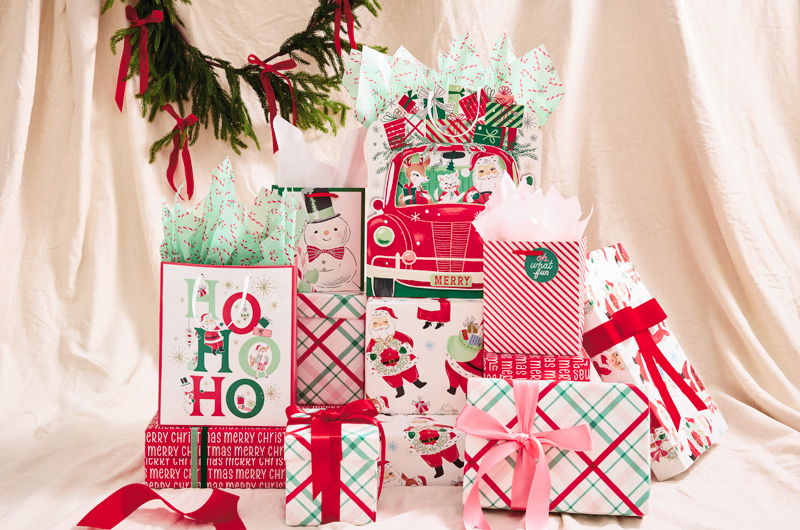 A collection of gifts wrapped, boxed and bagged in vintage-inspired gift wrap; the gifts sit on a cream-colored backdrop hung with a simple green garland with red velvet ribbon.