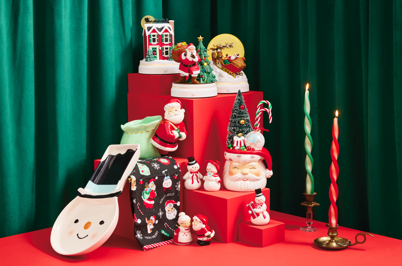 A collection of vintage-inspired decor and gifts sits on a red table top with a stair-step riser against a deep green velvet curtain background; the decor and gift items include a serving platter shaped like a snowman wearing a stove-top hat; a 3-piece set of figurines with light and sound that depict the events of the classic Christmas poem, 