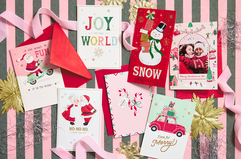 A collection of vintage-inspired Christmas cards lays on a pink and gray striped surface with pink velvet ribbon, silver tinsel and gold metallic ornaments strewn around them.