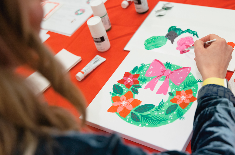 A woman paints a whimsical Christmas wreath on a piece of stretched canvas at a Canvas and Clinks Create & Learn Workshop at the Hallmark Christmas Experience.