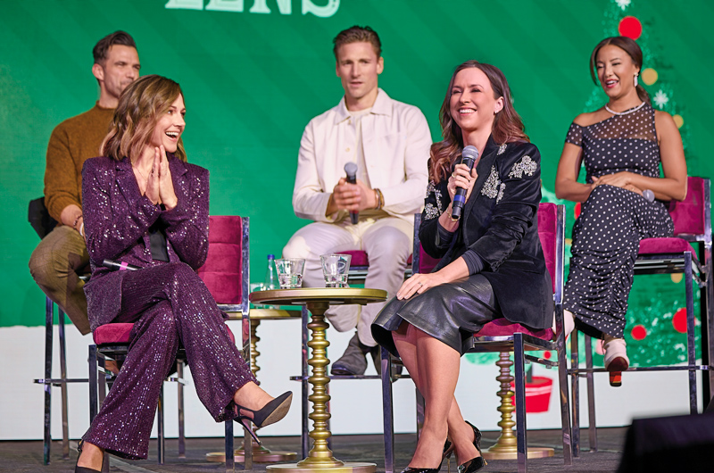 A group of five Hallmark movie stars, including Lacey Chabert, Andrew Walker, Benjamin Ayres, and Nikki DeLoach sit on stage at the 2024 Hallmark Christmas Experience during a Q&A panel.