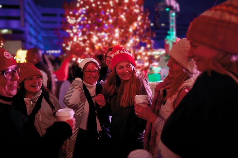 Attendees at the 2024 Hallmark Christmas Experience hold warm drink cups with lids and wear cozy knit caps as they're gathered in Crown Square, talking and laughing.
