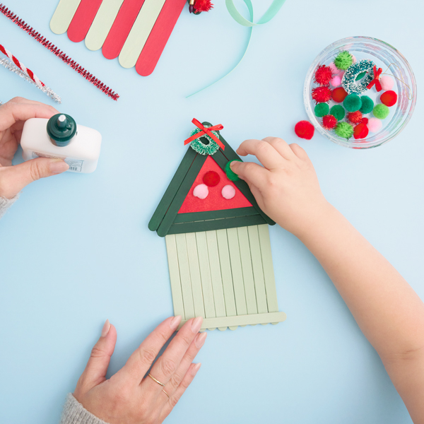 A child reaches into frame to press a fuzzy green pompom onto a dot of glue that her mother has applied to a wooden popsicle stick Christmas house facade.