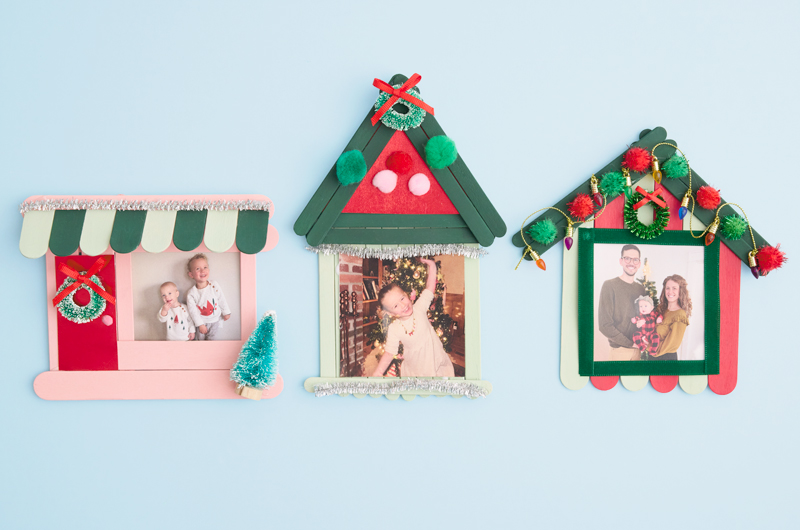 Three DIY photo frames made of wooden popsicle sticks, shaped and painted like houses decorated for Christmas, display photos of children and family.