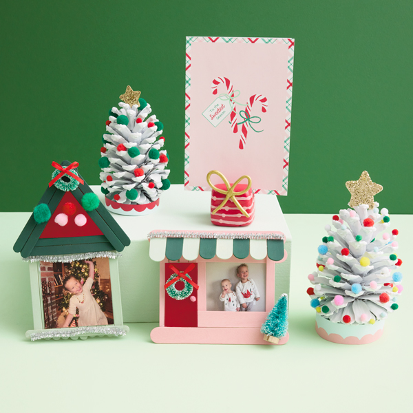 A trio of kids Christmas crafts, including Christmas card display stands made of air-dry modeling clay; a mini Christmas tree made from a painted pinecone and decorated with glitter and mini pompoms; and DIY mini photo frames made of wooden popsicle sticks shaped and decorated to look like houses at Christmas time.