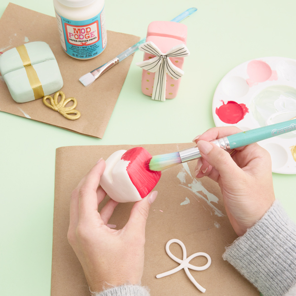 A woman paints a piece of white air-dry modeling clay with a bright, Christmassy red color of acrylic craft paint.