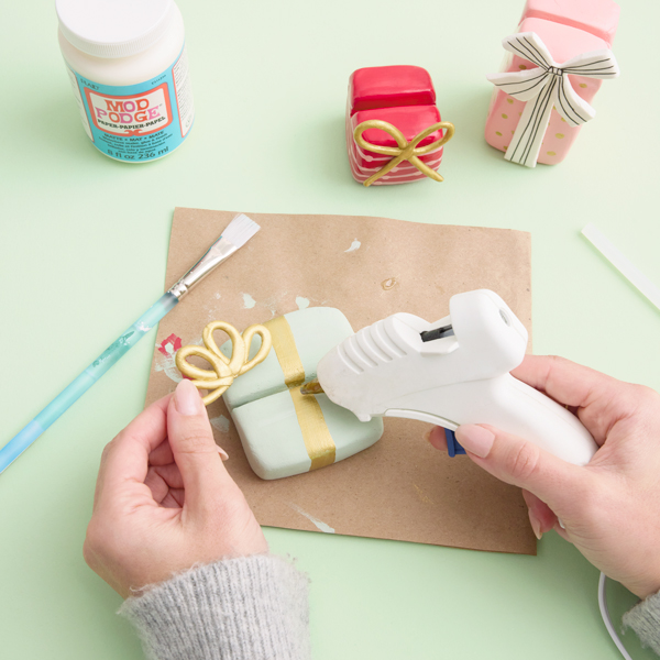 A woman hot glues a bow made from air-dry modeling clay and painted gold onto a base of similarly painted clay.