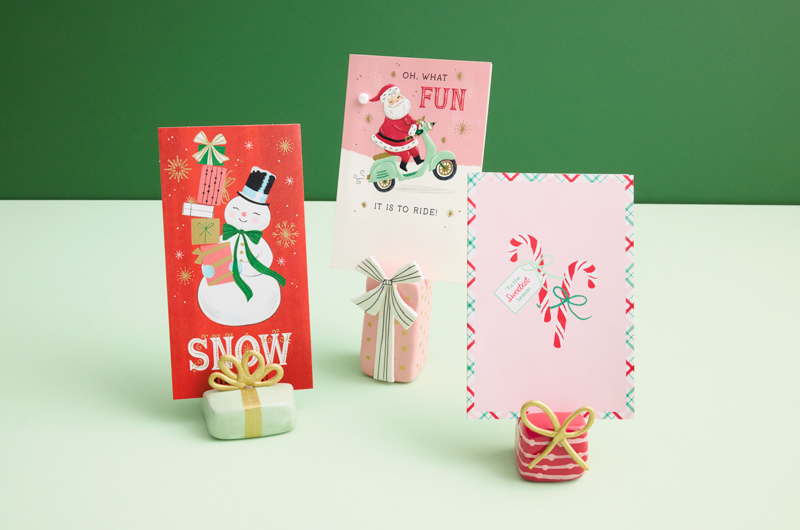 A trio of Christmas card display stands made of air-dried clay, shaped and painted to look like wrapped Christmas gifts, sits on a pastel green surface; each display stand holds a different Christmas card.