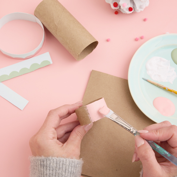 A woman's hands paint a piece of cardboard that has been cut from an empty toilet paper roll tube; she is painting it a pastel pink color.