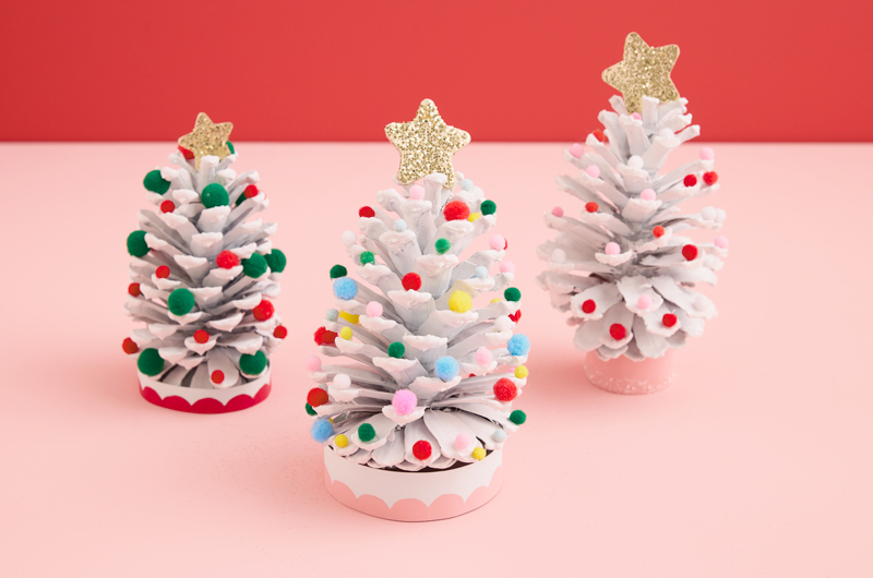 Three mini Christmas trees made from painted pine cones and decorated with miniature craft pompoms and gold glitter stars sit on a pastel pink surface.
