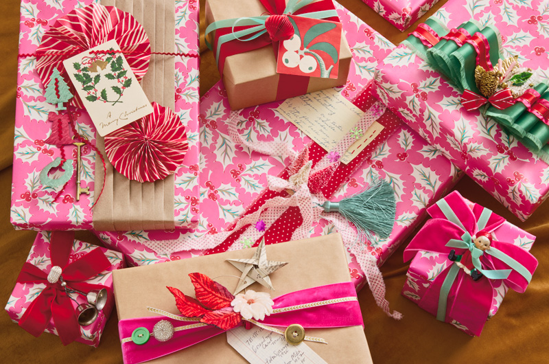 A pile of Christmas gifts wrapped in bright pink gift wrap that's covered in a holly pattern; the gifts are embellished with an assortment of ribbons, folded paper decorations, vintage items like skeleton keys, buttons, old recipe cards, etc.