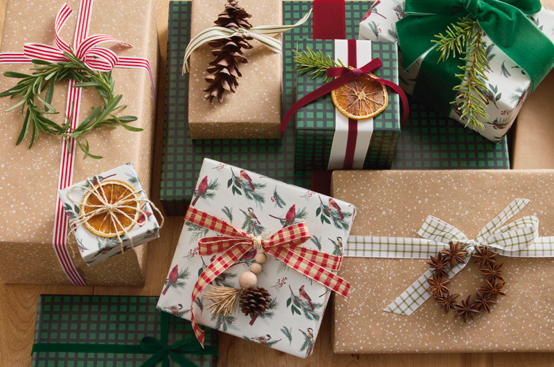 A pile of Christmas gifts wrapped in nature-inspired wrap in shades of kraft brown, dark green plaid, and cardinals and chickadees with greenery; the gifts are embellished with simple ribbon in check and solid colors, as well as natural items like pine cone, dried orange slices and sprigs of rosemary.
