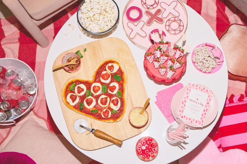 A table set up for a Secret Valentine party, including a heart-shaped pizza, white chocolate-covered strawberries, white chocolate-covered pretzels, pink and white paper straws, pink plates and napkins, as well as popcorn and candy; the table is surrounded by pink and red pillows and blankets; nearby is a white tub filled with ice and beverages in pink and red cans.