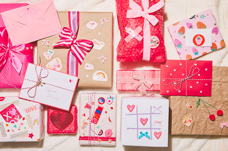 A collection of gifts at a Secret Valentine party, decorated with different kinds of wrap in shades of pink, red, white and kraft paper brown, wrapped with ribbons of different widths and materials in pink, red and white patterns, and covered in fun Valentine's Day-themed stickers.