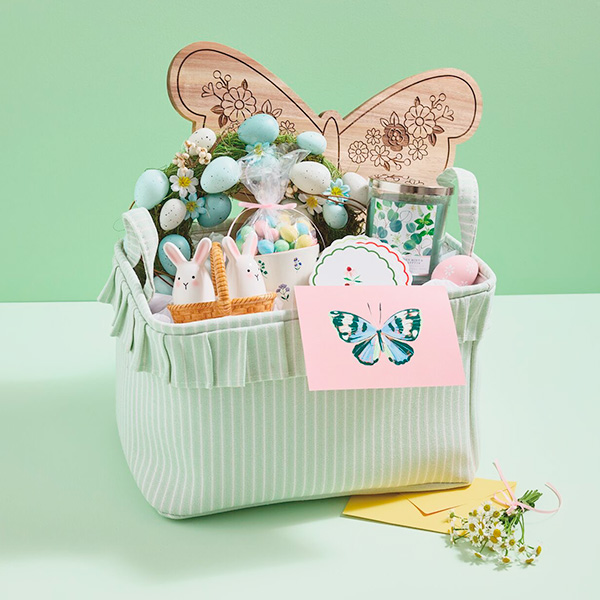 An Easter basket idea for an adult that's spring decor themed; the Easter basket is filled with gifts including a butterfly-shaped charcuterie board, a floral mug, a set of ceramic salt and pepper shakers in the shape of white bunnies, a decorative wreath covered in pastel eggs and dainty florals, a set of four floral coasters, a eucalyptus-scented candle, and a card with a butterfly on the front.