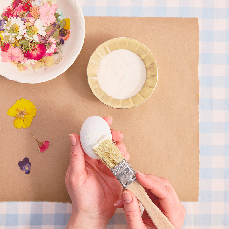 A woman holds a white hardboiled egg in her left hand while painting white glue onto the shell with her right hand; nearby are a bowl of glue and a bowl of pressed, dried flowers.
