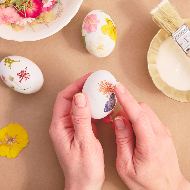 A woman applies pressed, dried flowers to the glue she's painted on to the shell of a hardboiled egg; nearby are eggs she has already finished decorating in a similar fashion.