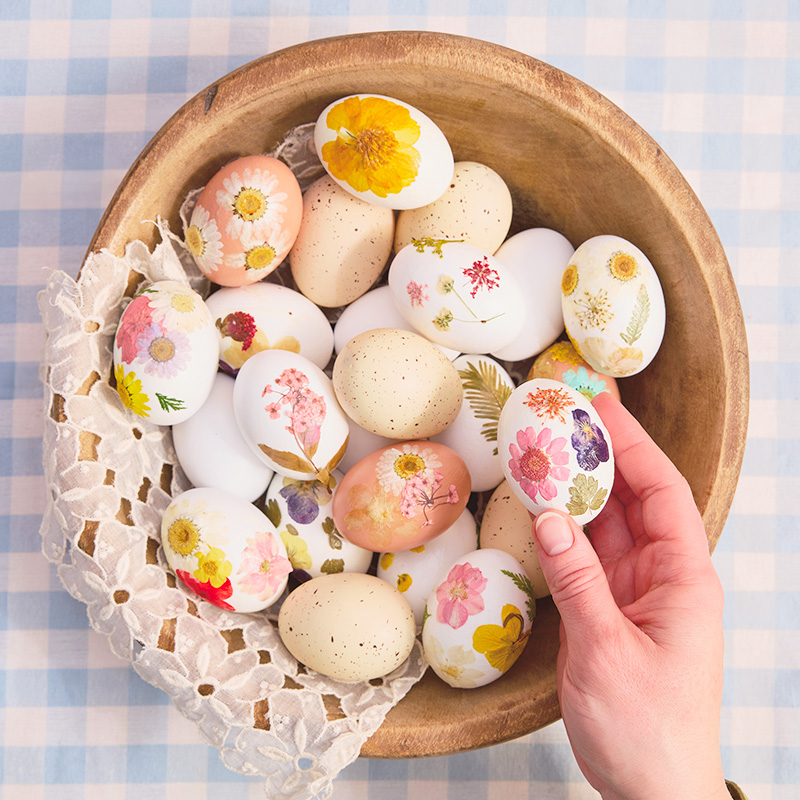 A woman's hand reaches into a wooden bowl to place an egg inside it; the egg has been decorated with pressed, dried flowers, as have many other eggs already resting in the bowl.