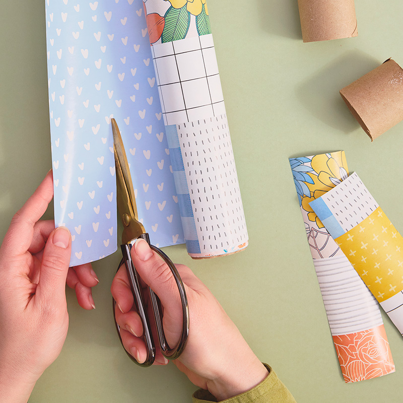 A woman's hands are shown cutting wrapping paper into two-inch strips that will be used to cover the cardboard tube sections laying nearby.