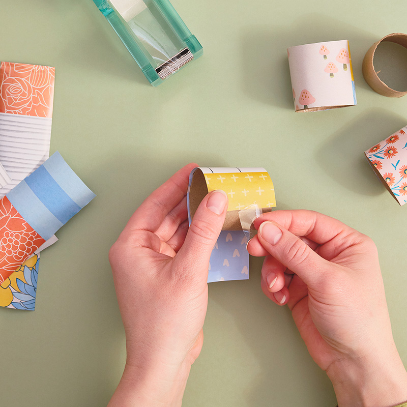 A woman covers cardboard tubing with a section of wrapping paper of the same two-inch width, taping it in place.