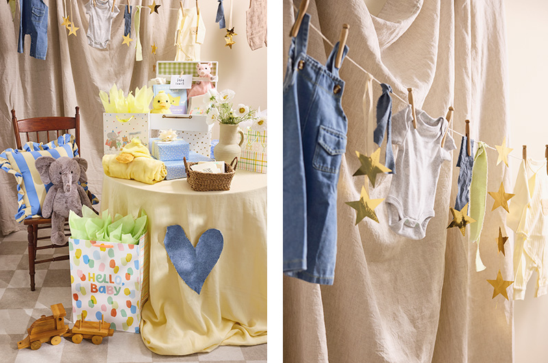 A baby shower decor idea that involves thrifted or second-hand baby clothes pinned up on a clothes line with antique-style wooden clothes pins to create a cute, clothing-themed bunting; the clothing would be taken down after the shower and given to the expectant mom; next to a closeup of the bunting idea is a gift table decoration idea, with a light yellow blanket covering the table and a blue heart cut out of old denim pinned to it, decorating the front of the gift table cloth; sitting on top of the table are various wrapped and bagged baby shower gifts, along with a basket full of greeting cards and a ceramic jug full of flowers; nearby is a chair with a light yellow and blue striped pillow and a plush elephant toy sitting on it.