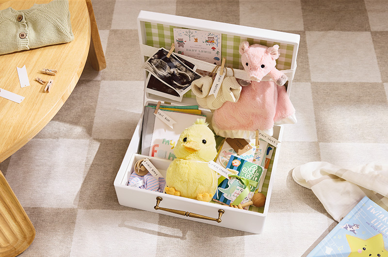 A baby shower gift idea comprised of a memory box; inside are items marked with handmade tags, such as a plush duck with a tag that reads, 