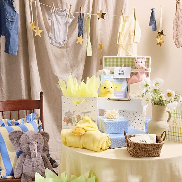 A baby shower decor idea that involves thrifted or second-hand baby clothes pinned up on a clothes line with antique-style wooden clothes pins to create a cute, clothing-themed bunting; the clothing would be taken down after the shower and given to the expectant mom; next to a closeup of the bunting idea is a gift table decoration idea, with a light yellow blanket covering the table and a blue heart cut out of old denim pinned to it, decorating the front of the gift table cloth; sitting on top of the table are various wrapped and bagged baby shower gifts, along with a basket full of greeting cards and a ceramic jug full of flowers; nearby is a chair with a light yellow and blue striped pillow and a plush elephant toy sitting on it.