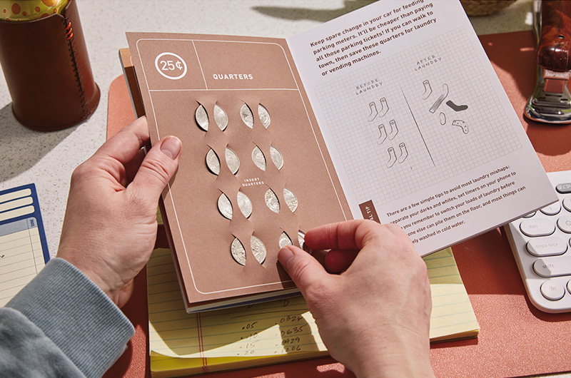 An inside spread of the Graduation Survival Guide book, with slots to place eight quarters on the lefthand page; on the righthand page are tips for spending those quarters on parking meters and laundry machines.