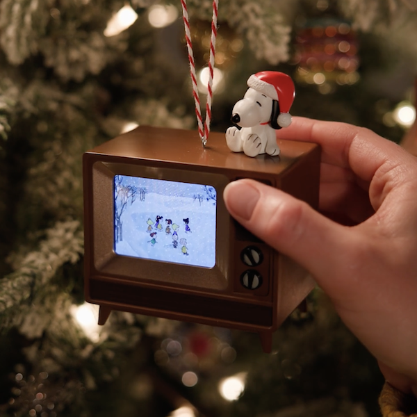 A hand reaches into frame to press the button that activates a Keepsake Ornament in the form of a tiny console TV with a Santa hat-wearing Snoopy sitting on top of it; the tiny television actually plays a video of the opening scenes from the movie, 