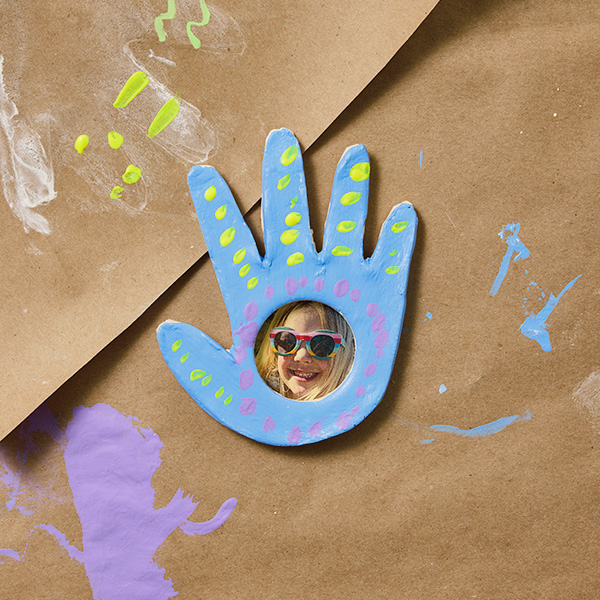 A photo frame magnet made out of air-dry clay and in the shape of a child's hand sits on a piece of brown kraft paper smeared here and there with paint; the handprint photo frame has been painted blue and decorated with yellow and purple dots; in the palm of the hand is a perfect circle cut out that reveals a child's picture.
