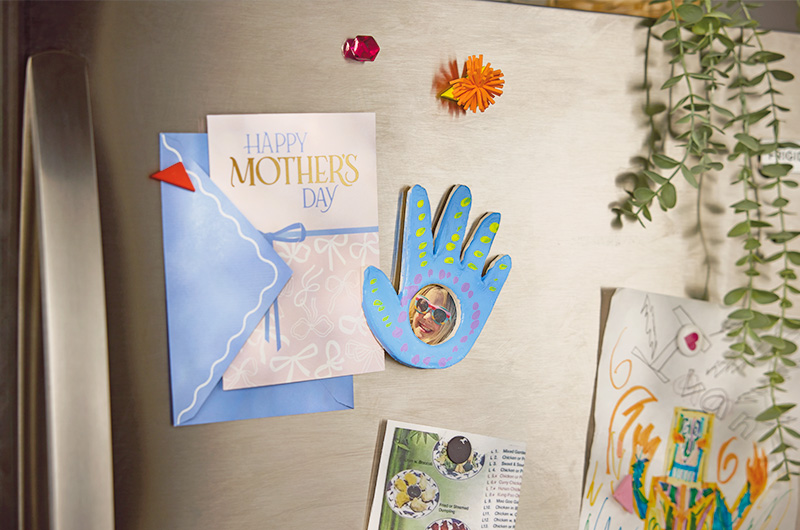 A photo frame magnet made out of air-dry clay and in the shape of a child's hand is stuck to the front of a refrigerator; the handprint photo frame has been painted blue and decorated with yellow and purple dots; in the palm of the hand is a perfect circle cut out that reveals a child's picture; the handprint photo frame magnet is helping to hold a Mother's Day card up on the refrigerator door; additional drawings, a takeout menu and magnets are also stuck to the refrigerator door.