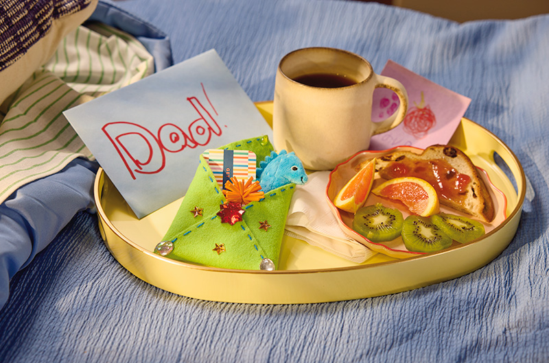 A breakfast tray made up for a dad by his children for Father's Day; the tray holds a mug filled with coffee, a plate with toast and jam, as well as orange and kiwi slices; a greeting card envelope with 
