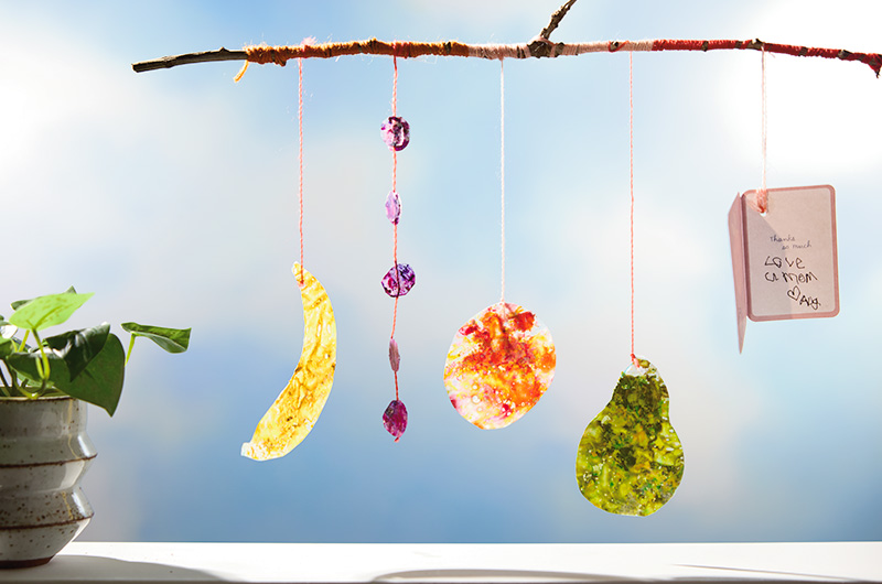A DIY suncatcher made by a child as a gift to their mom for Mother's Day; the suncatcher is comprised of a stick wrapped with colorful string, with pendants hanging from it; the pendants are in different shapes and are made from various colors of melted wax crayon shavings; at the end of one of the strings hangs a mini greeting card and inside the card in childish scrawl is written, 