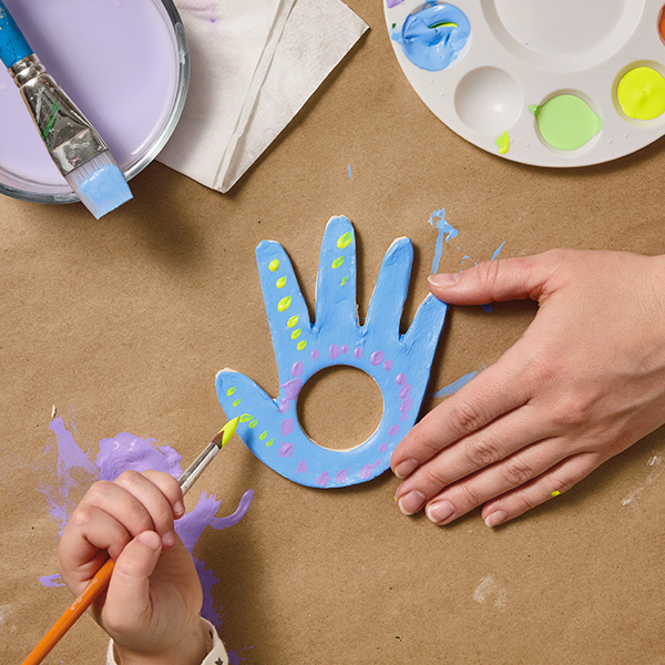 A woman's hand reaches into frame to hold a dried piece of air-dry clay that's been traced and cut out into the shape of a child's hand; there is a perfect circle cut out in the palm of the clay hand; the hand of the child that the craft is modeled after is reaching into frame with a paintbrush to decorate the piece; nearby is a small plastic paint palette with different colors of paint on it.