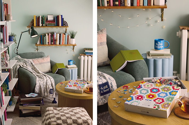 A vignette setting showing what a book nook-style calming corner for adults could look like, with multiple shelves of books strung with battery-powered fairy lights; a few potted, live plants; a table for assembling puzzles and drinking tea; a cozy, overstuffed-style chair with a throw blanket draped over it and a task lamp positioned above it for reading; and a side-table with a stack of books topped by a mug.