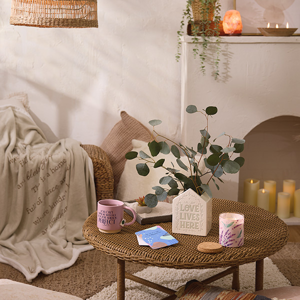 A vignette setting showing what a spa-like calming corner for adults could look like, with wicker furniture, live plants, soothing neutral colors, candles, soft blankets and comforting items like an aroma therapy candle, a mug of tea and a mini zen garden.