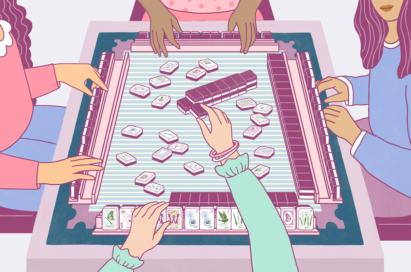 An illustration of four women playing at a mahjong table; the illustration is from the perspective of the woman who is currently discarding a tile; her friends look on as she discards; the tile set resembles the set created by Hallmark in partnership with mahjong tile set brand Miss Mahjong.