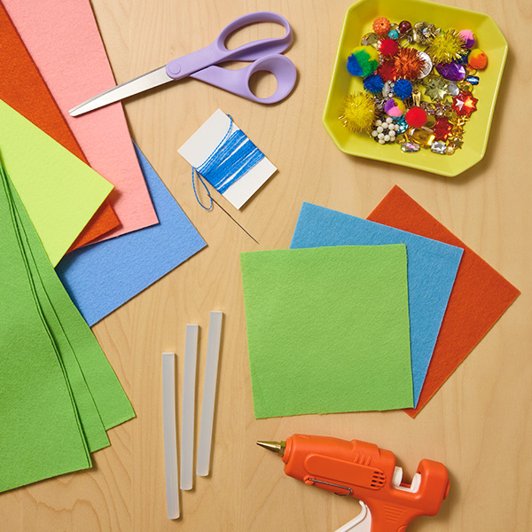 Materials needed to create the Felt Gift Envelope include a sheet of felt fabric (also known as crafting felt) in any color, scissors, embroidery floss, embroidery needle, hot glue gun, and embellishments like gems, buttons, and mini pom-poms.