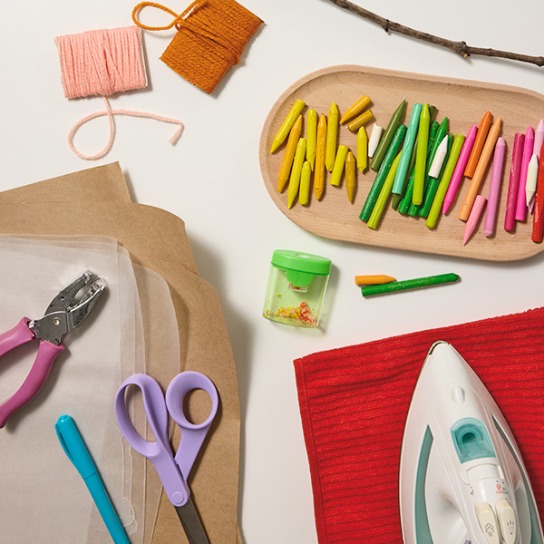 Materials needed to create the melted crayon suncatcher craft include a small, thin stick; at least two different colors of yarn or twine; crayons in a variety of bright colors; a pencil sharpener; a hole punch; a pair of scissors; a marker; pieces of wax paper; pieces of brown craft paper; a kitchen towel; and a clothing iron.